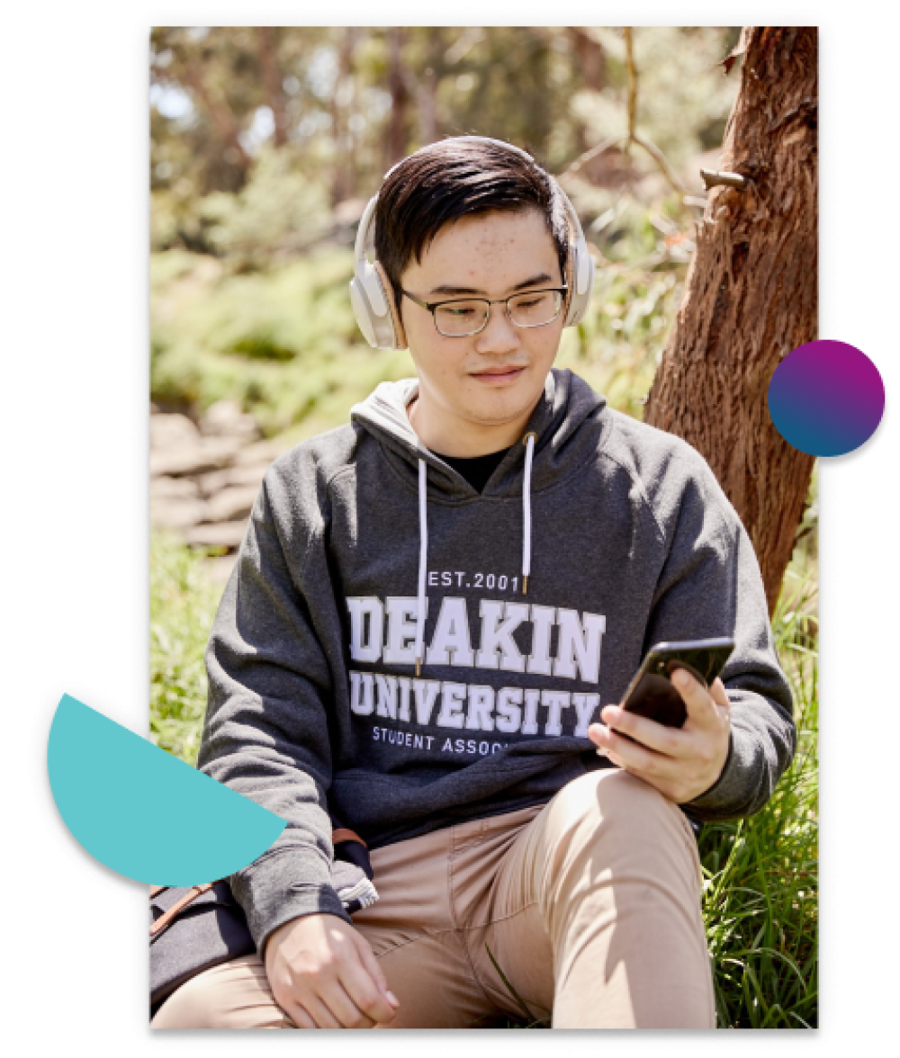 Male student wearing a DUSA Hoodie sitting outside on the grass wearing headphones and looking at his mobile phone