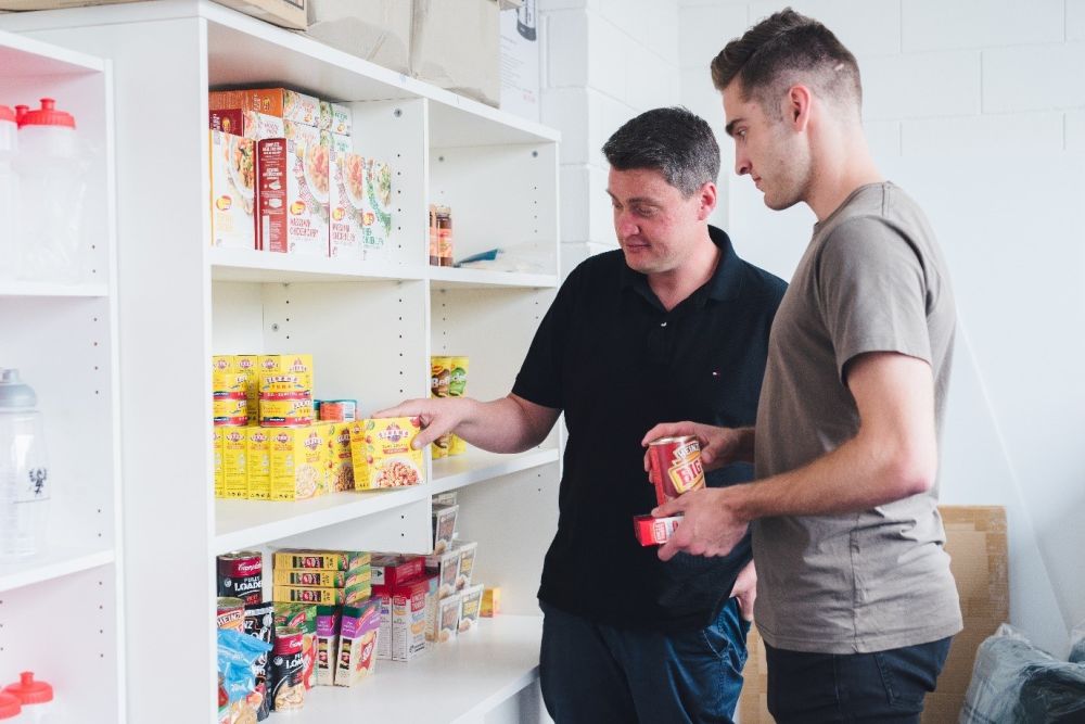 Male student and Advocacy staff member standing in front of shelves of food at the DUSA Survival Centre