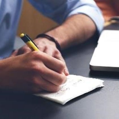 Male sitting at a desk in front of a laptop writing notes