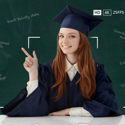 Female student wearing a graduation cap and gown sitting at a desk in front of a black board