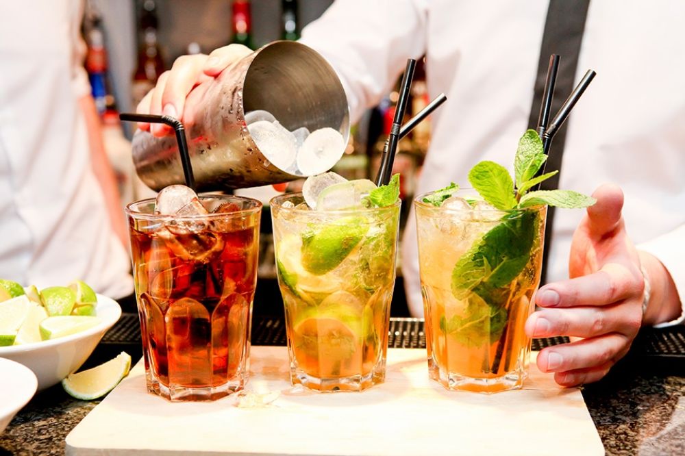 A person pouring ice cubes into three tall glasses filled with liquid to make cocktails