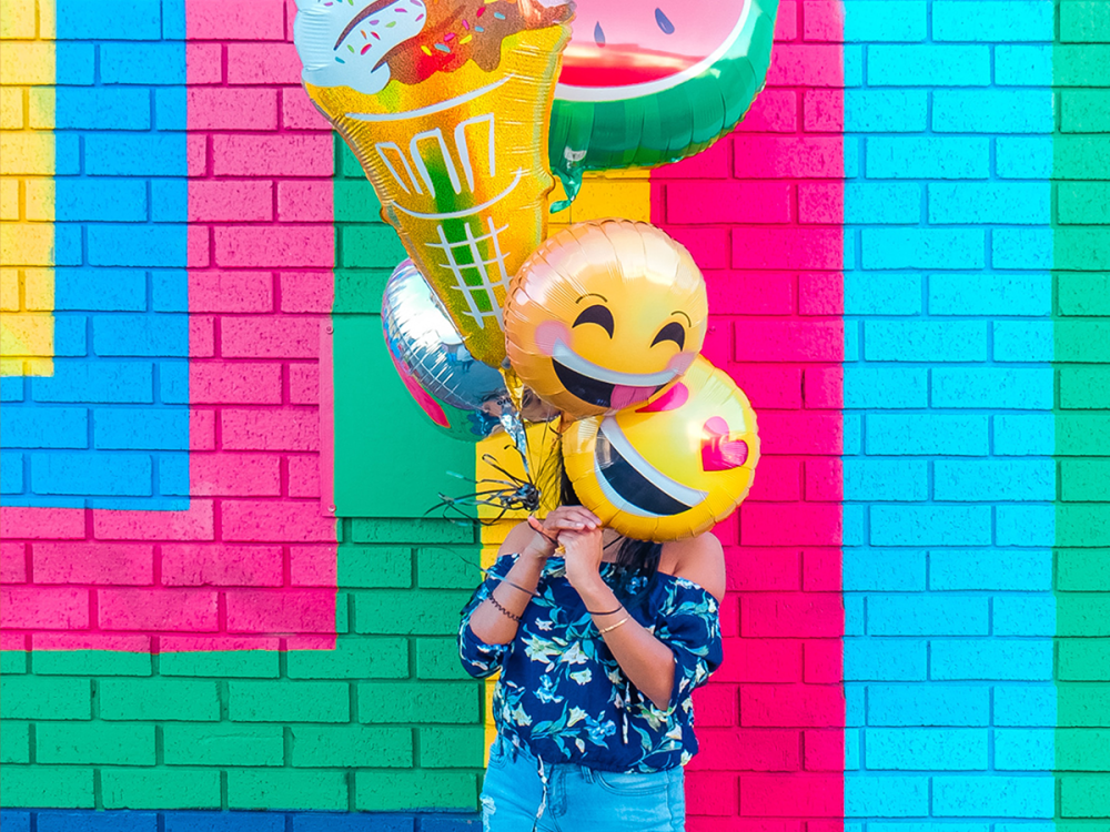 Person standing in front of a bright coloured wall with bright coloured balloons in front of their face.