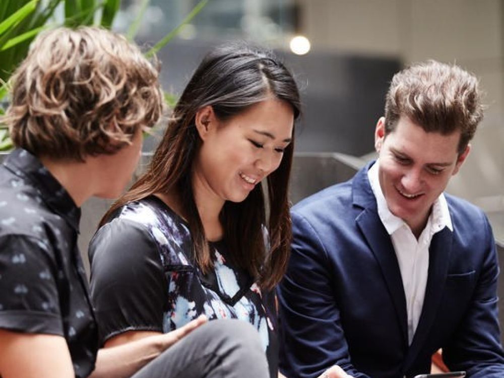 3 people sitting next to each other, left to right: male, female, male. Looking at female's tablet screen.