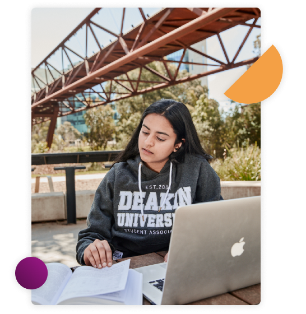 Female student sitting at an outdoor table looking through her textbook with her laptop open